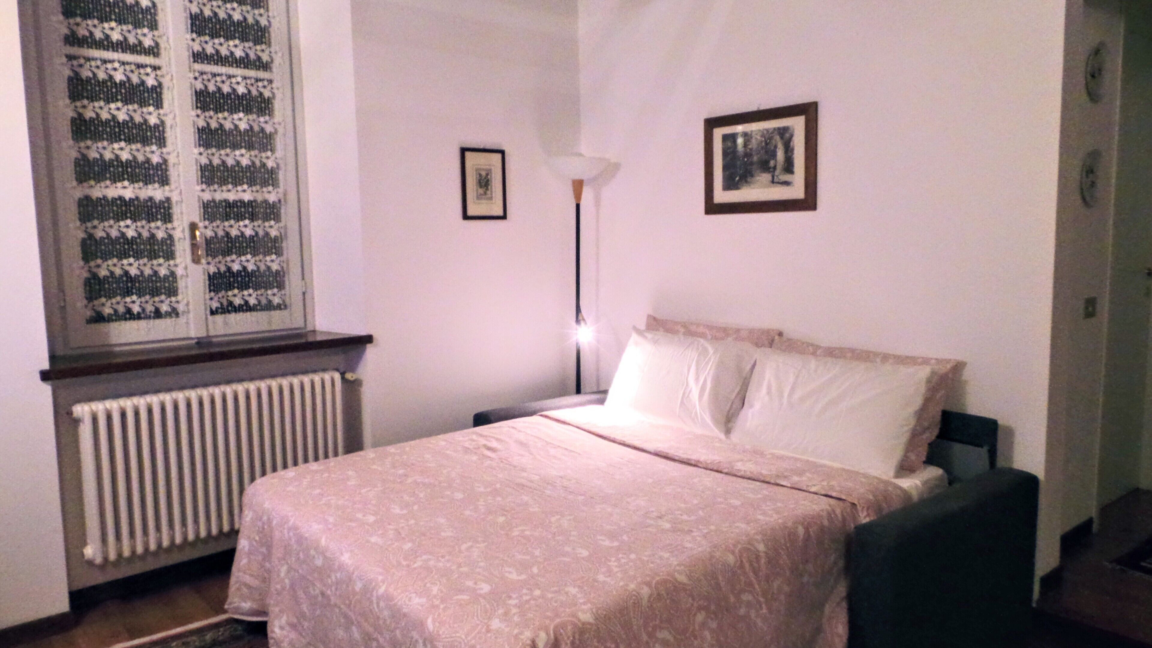 A bedroom with a bed, a radiator, a window with a patterned blind, a floor lamp, and two framed pictures on the wall.