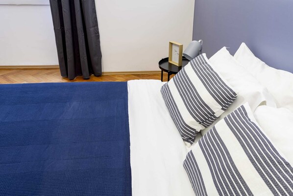 A neatly made bed with striped pillows and a blue rug, a small table with a picture frame, and a grey wall.