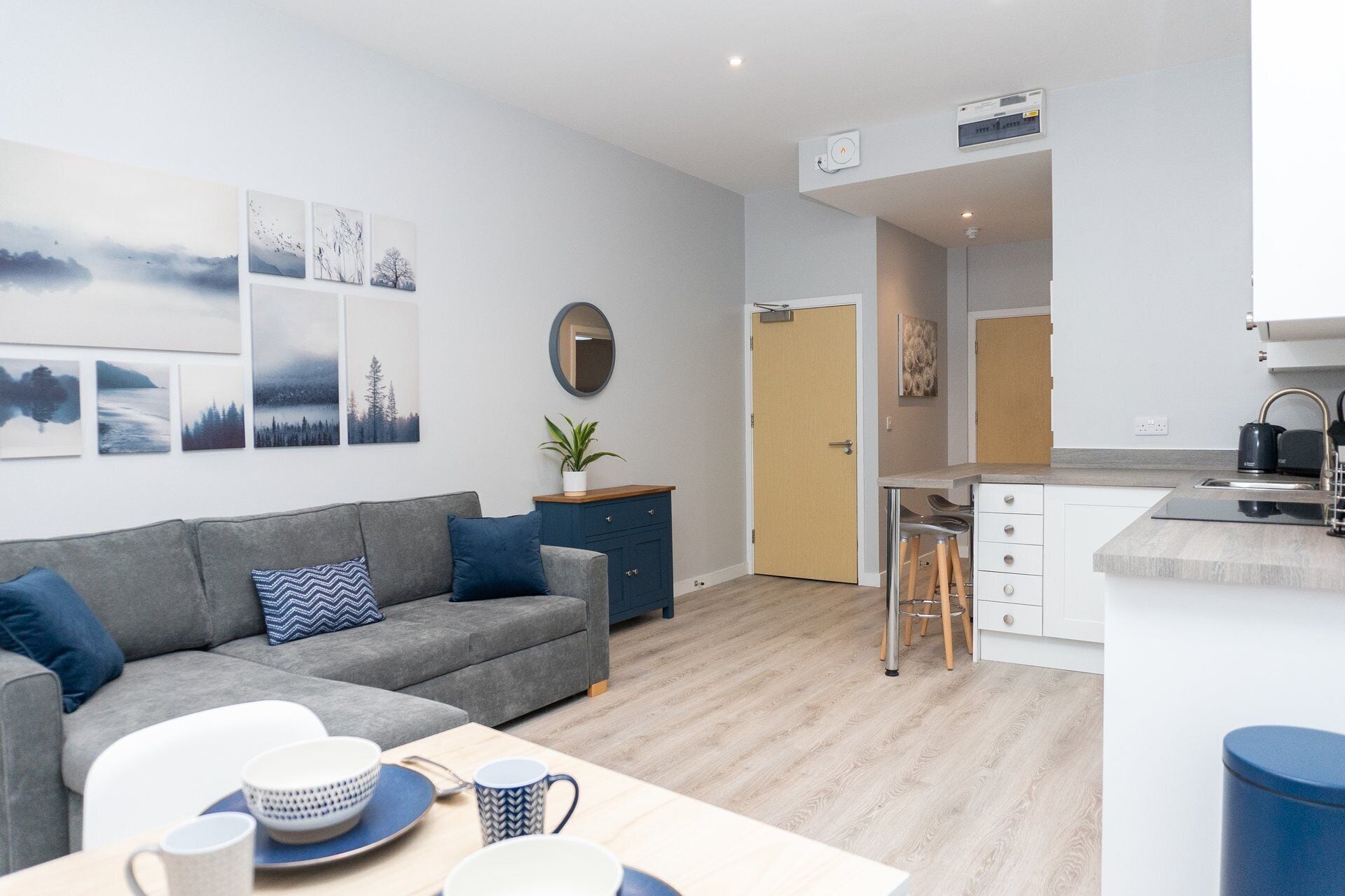 Una sala de estar moderna con un sofá gris, una mesa de centro con vajilla, y un área de cocina con una encimera y taburetes.