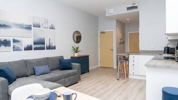 Una sala de estar moderna con un sofá gris, una mesa de centro con vajilla, y un área de cocina con una encimera y taburetes.