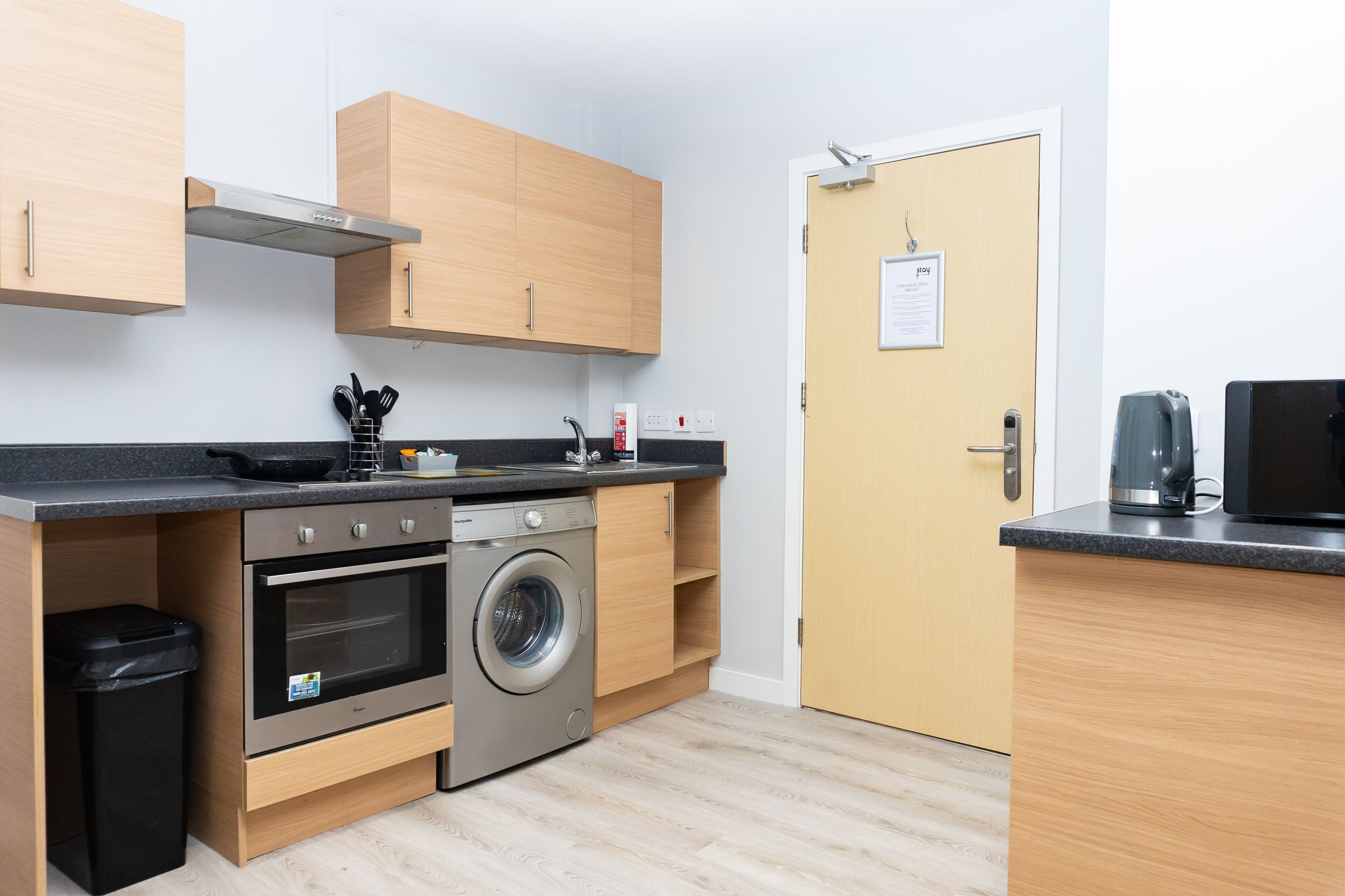 A compact kitchen with wooden cabinets, a built-in oven, a washing machine, and a coffee maker.