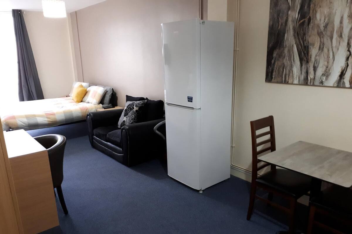 A compact hotel room with a bed, a sofa, a dining table, a refrigerator, and a chair.