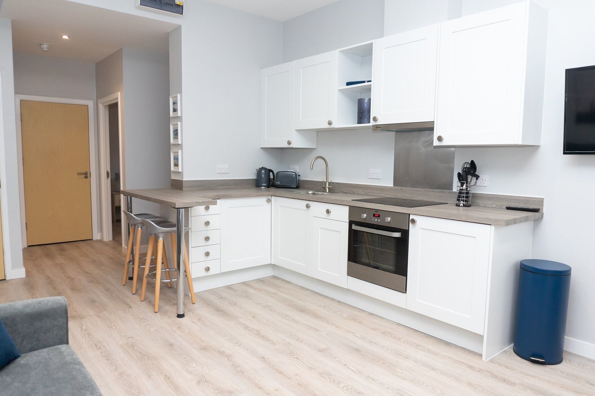 Una cucina moderna con mobili bianchi, forno integrato e lavandino. C'è uno sgabello da bar e un cestino.