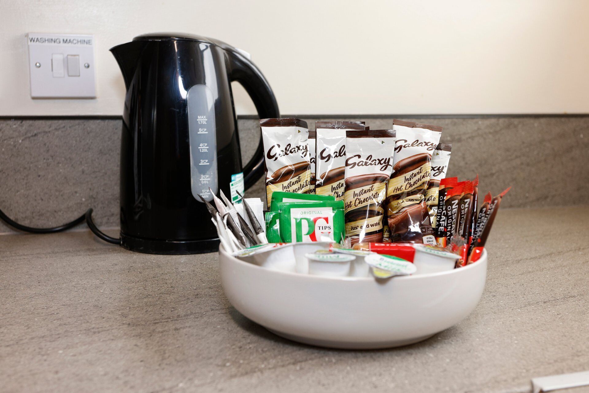A kitchen counter with a black kettle, a bowl of instant coffee packets, and a coffee grinder.
