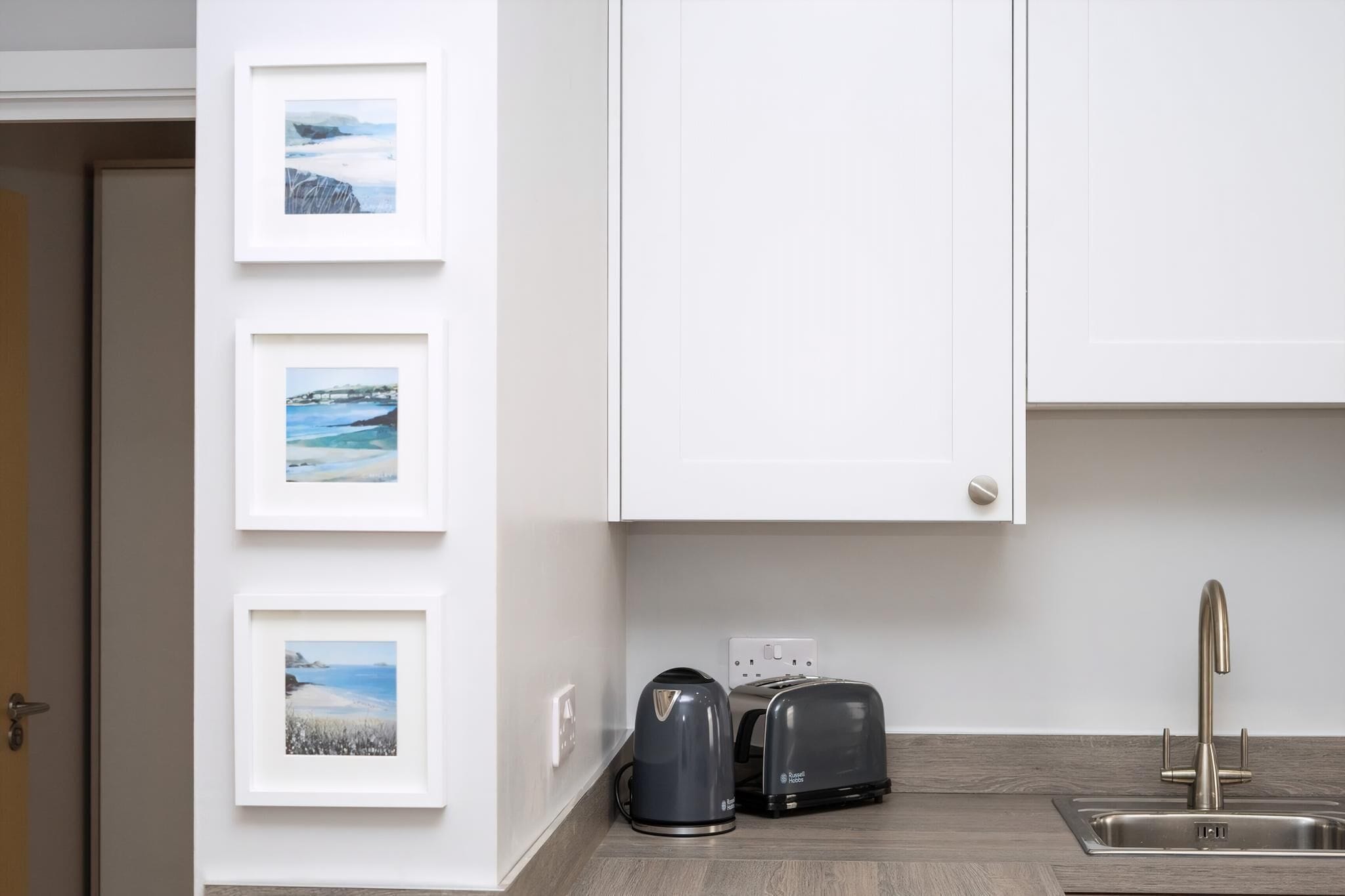 Een keuken met een moderne gootsteen, een broodrooster en een waterkoker. Er hangen drie ingekaderde foto's aan de muur.