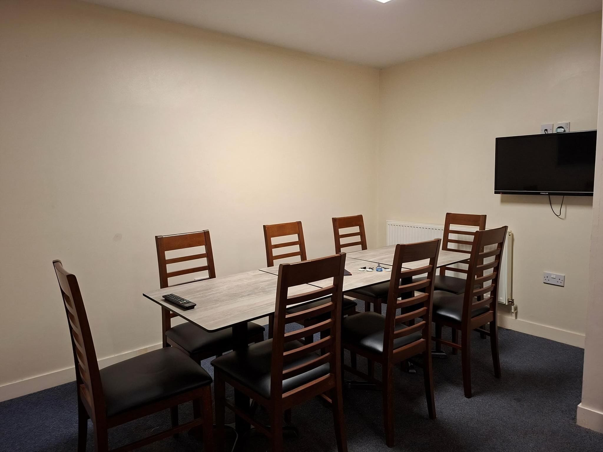 A room with a rectangular table, six wooden chairs, a flat-screen TV mounted on the wall, and a radiator.