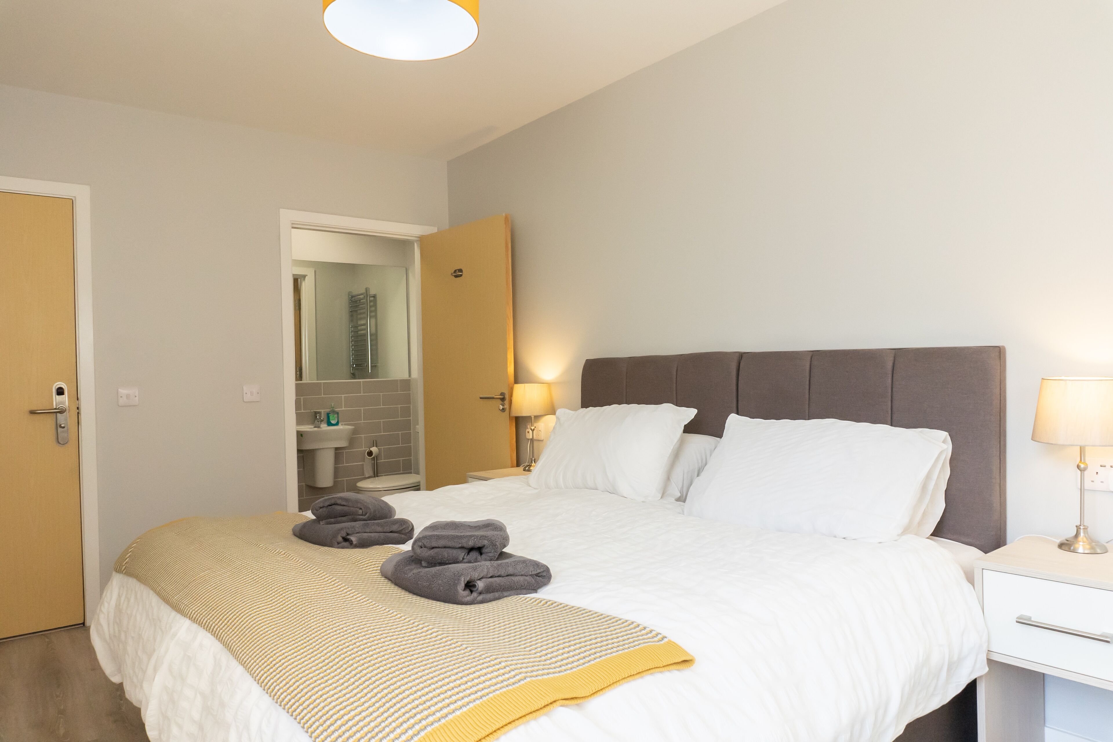 Una cama bien hecha con ropa de cama blanca y amarilla, una mesita de noche con una lámpara, y un baño visible a través de una puerta abierta.