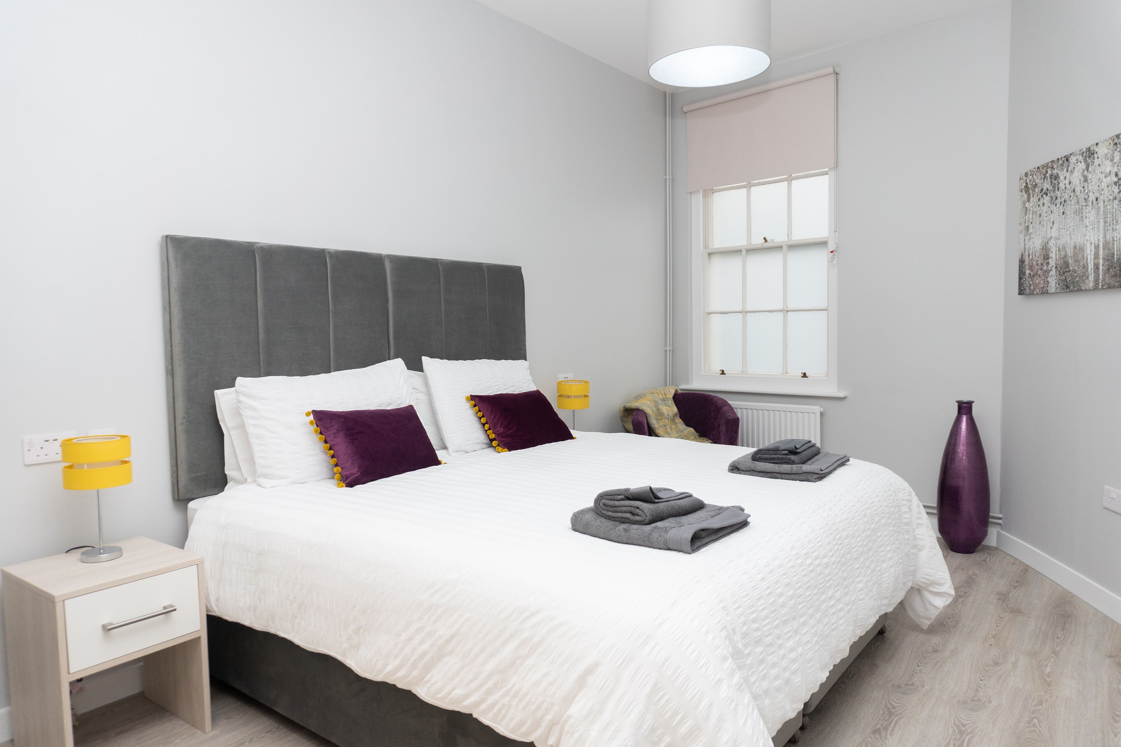 Een keurig opgemaakt bed met wit beddengoed en paarse kussens, een nachtkastje met een lamp, en een raam met zonwering.