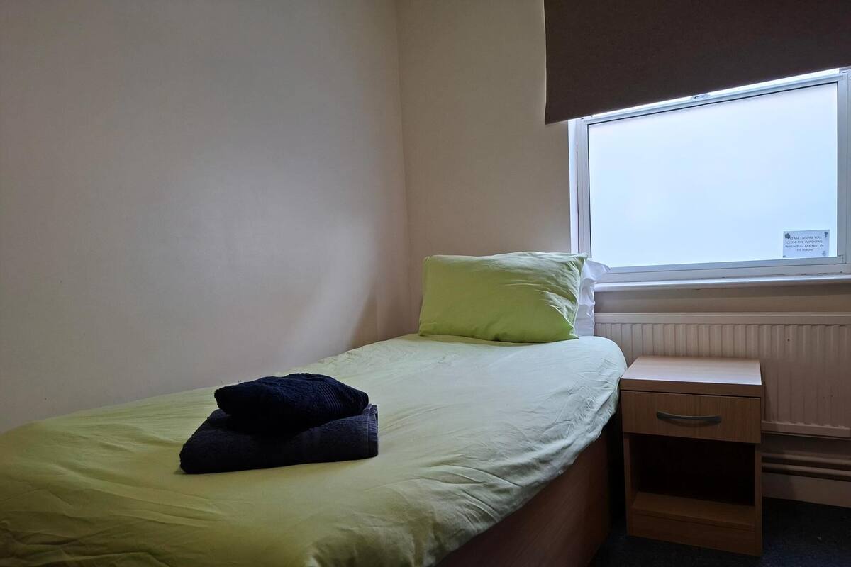 A single bed with a green bedspread and a dark pillow, a wooden nightstand with a drawer, and a window with a sign.