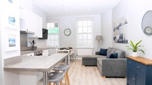 A modern kitchen with a central island, bar stools, a grey sofa, and a blue cabinet. - Spa Court (Cheltenham)