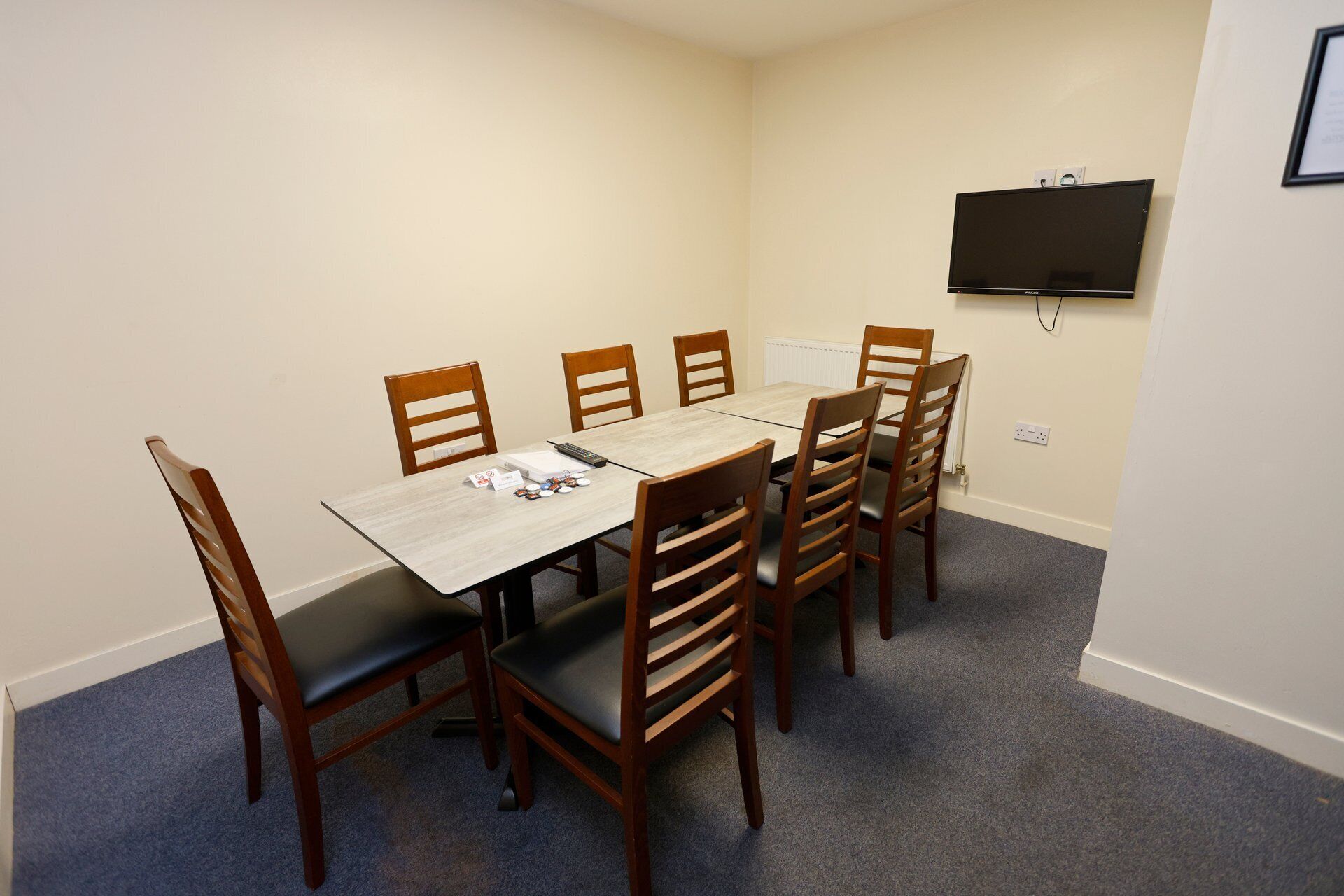 A room with a rectangular table, six wooden chairs, a flat-screen TV mounted on the wall, and a carpeted floor.