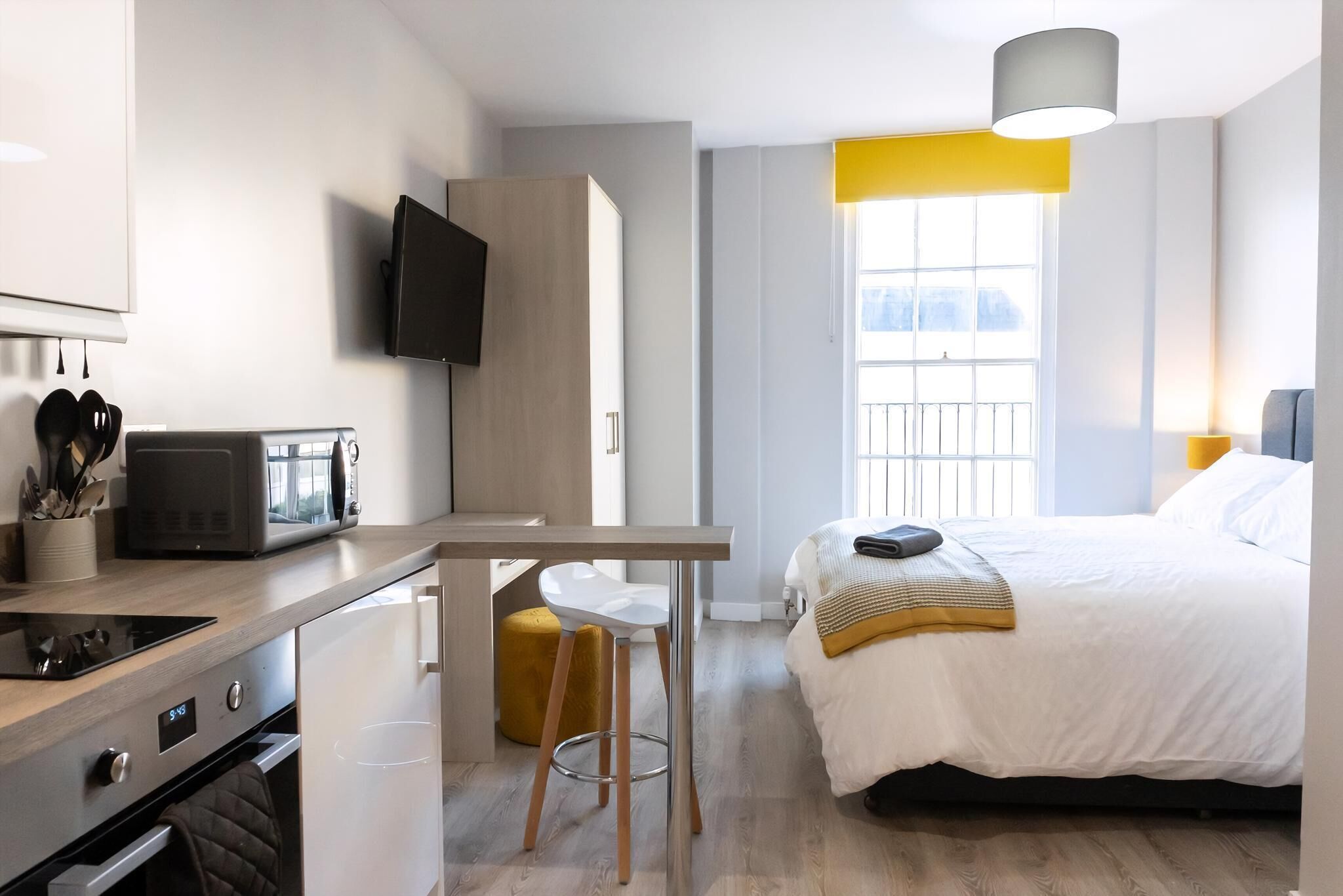 Una camera d'albergo moderna con angolo cottura, un letto con una coperta gialla, una TV a parete e una finestra con tenda gialla.