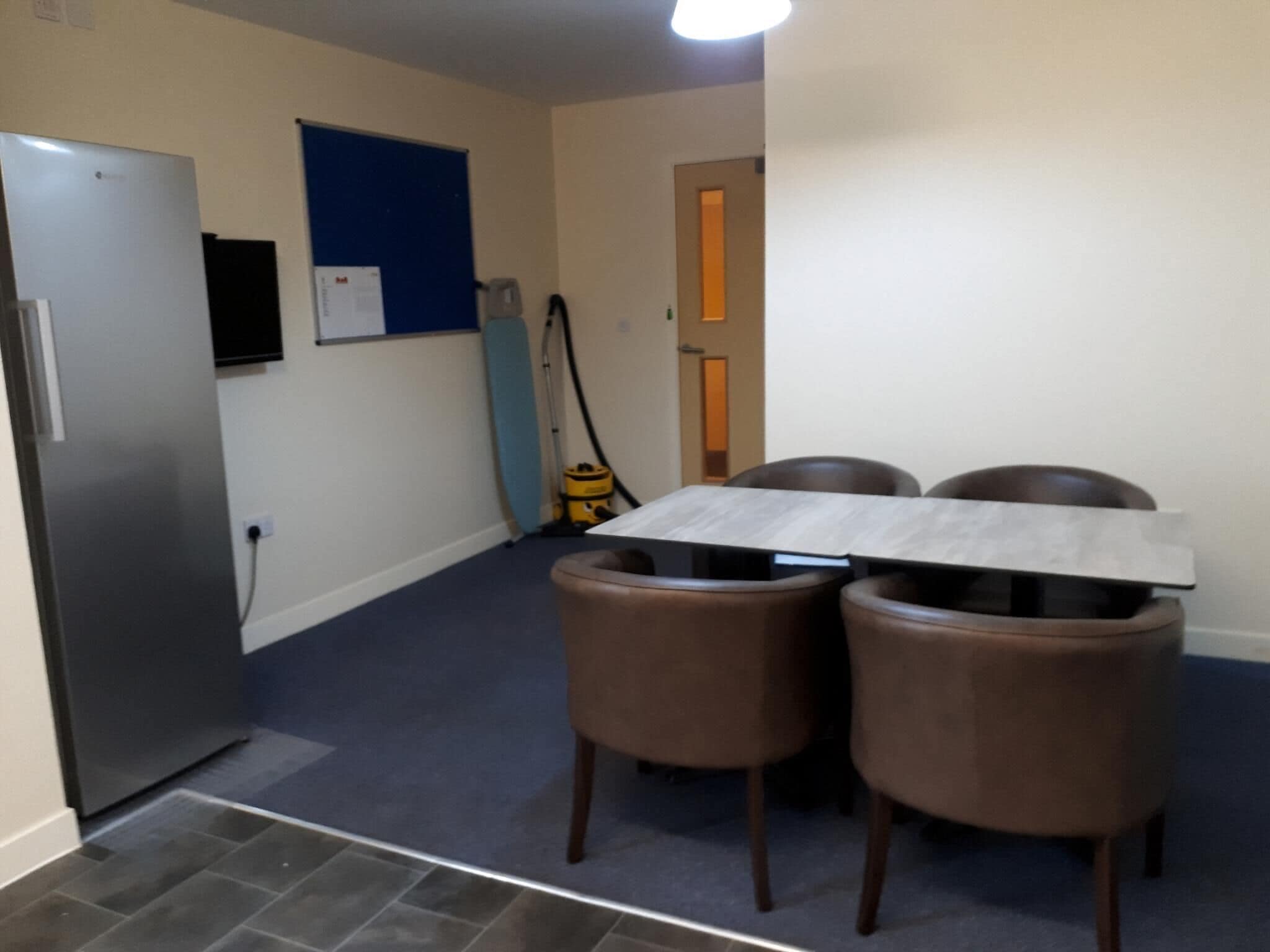 A compact kitchen with a refrigerator, a blue bulletin board, and a vacuum cleaner.