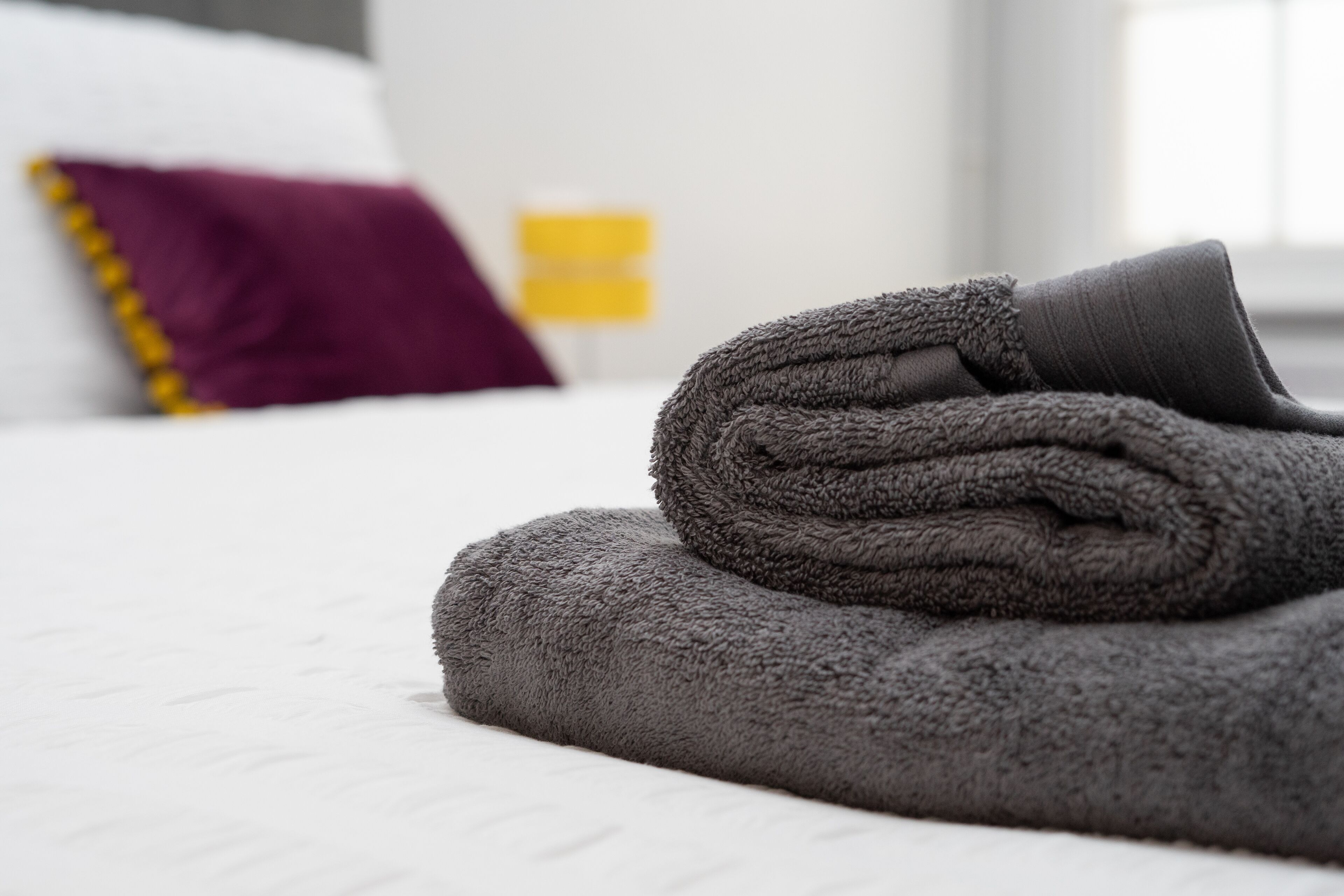 Una toalla gris doblada con cuidado sobre una cama con ropa blanca, un almohadón morado y un objeto amarillo al fondo.
