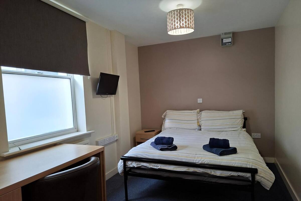 A bedroom with a bed, a desk, a chair, a TV, and a window.