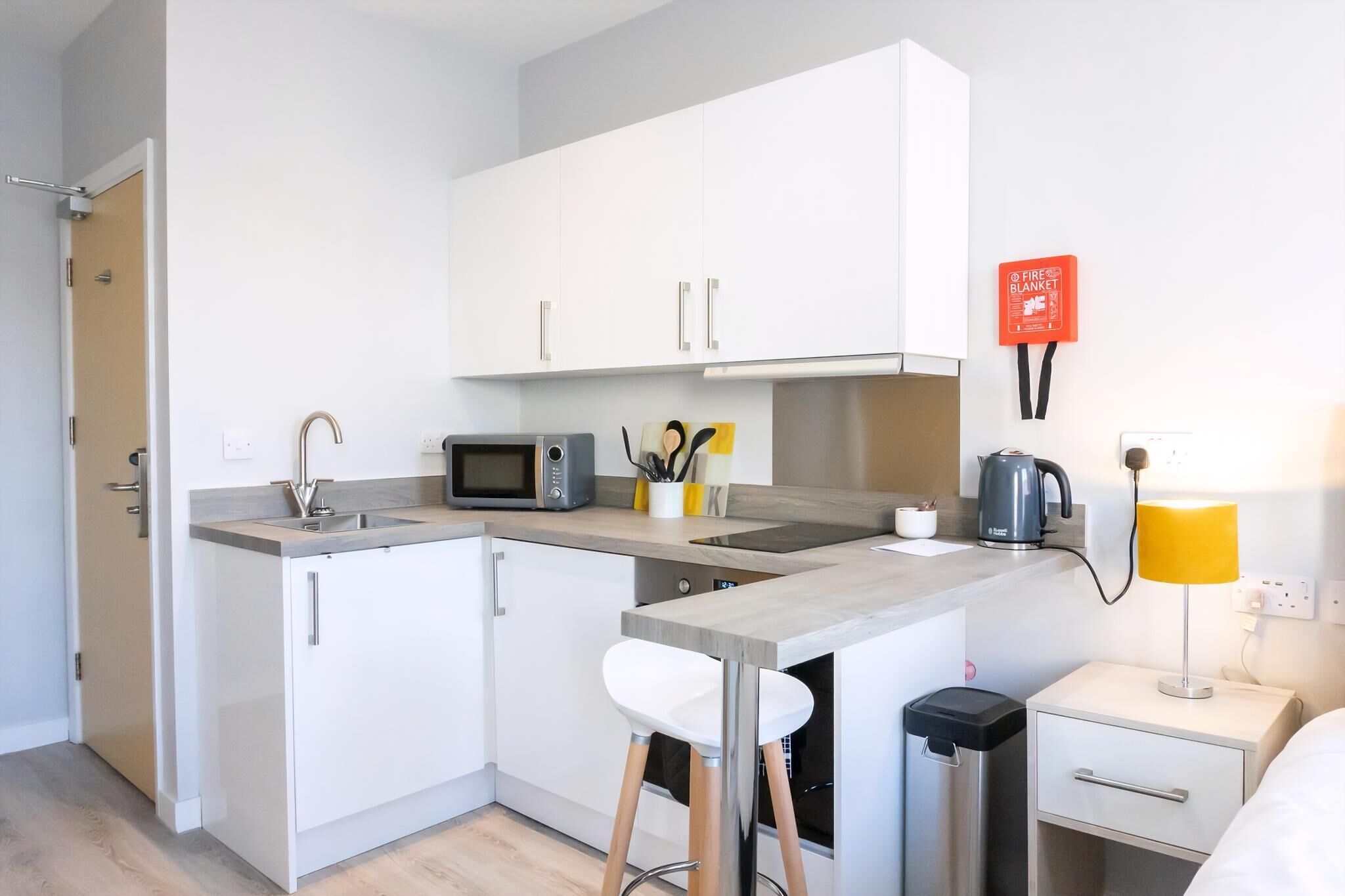Une cuisine compacte avec des meubles blancs, un four à micro-ondes, une bouilloire et un petit tabouret de bar.