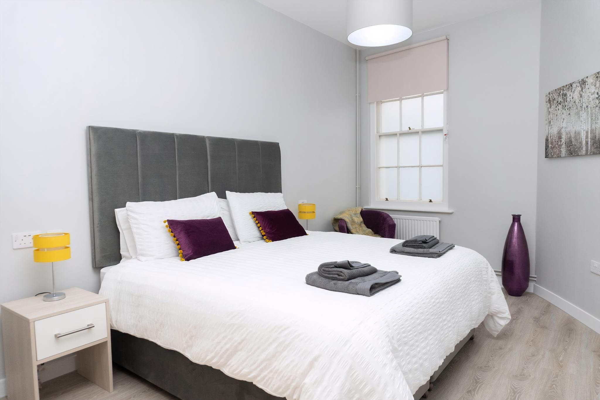 A modern bedroom with a large bed, a bedside table with a lamp, a window, and a decorative vase.