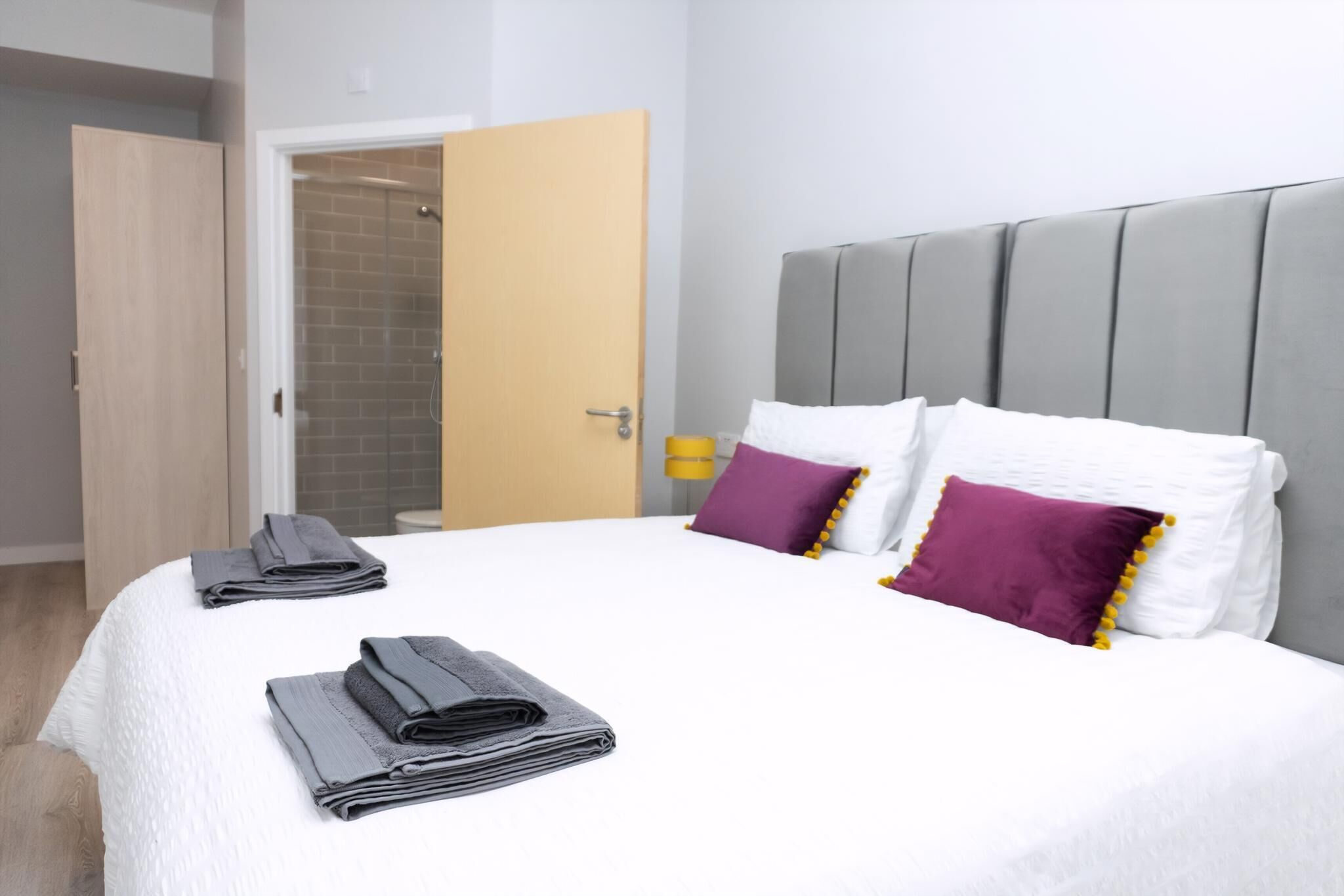 Una cama bien hecha con toallas dobladas, un cabecero gris y una puerta de madera que da al baño.