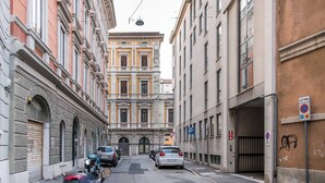 Una stretta via cittadina con auto e motociclette parcheggiate, edifici a architettura classica e un segnale di divieto di sosta.
