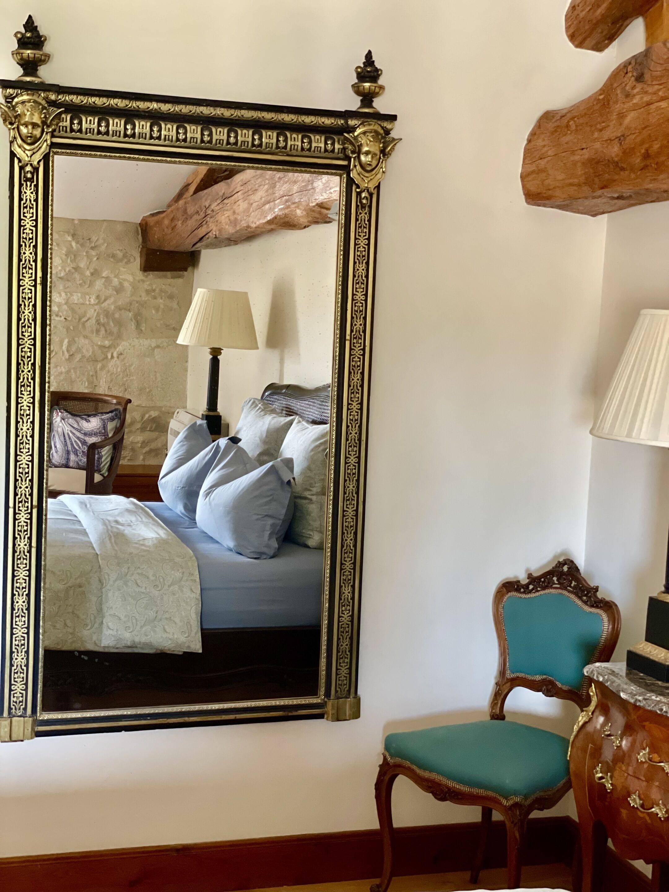 A bedroom with a large ornate mirror, a bed with multiple pillows, a nightstand, a lamp, and a wooden chair.