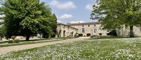 Un edificio storico in pietra con un giardino pieno di fiori bianchi e alberi grandi.