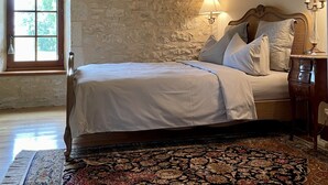 A bedroom with a stone wall, a wooden ceiling beam, a bed with white bedding, a patterned rug, and a nightstand with a lamp.