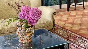 A room with a sofa, a side table with a vase of flowers, a decorative cabinet with intricate carvings, and a mural on the wall depicting a landscape.