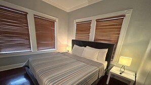 A bedroom with a large bed, two bedside tables, and three windows with blinds.