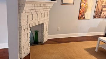 Una sala de estar con chimenea de ladrillo, un televisor encima, un sillón marrón y dos cuadros en la pared.
