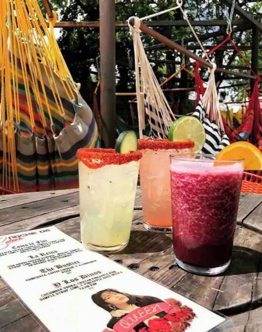 A wooden table with three drinks, a menu, and a hammock in the background.
