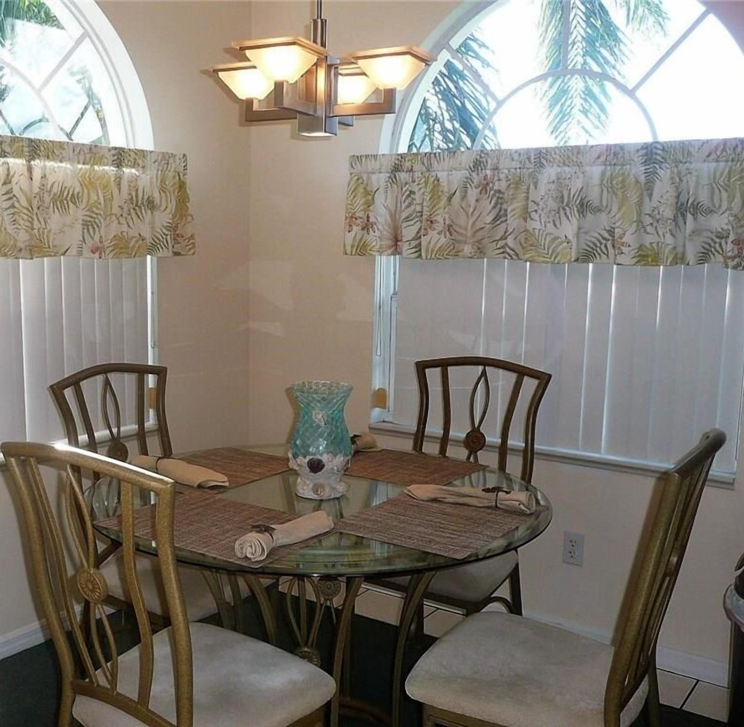 A dining area with a glass table surrounded by wooden chairs, a decorative vase, and a window with a view of palm trees.