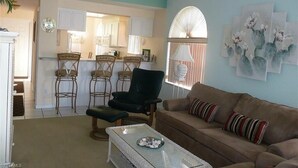 A living room with a brown sofa, a glass coffee table, and a black armchair. There is a kitchen area with white cabinets and a dining table with chairs. - Beautiful open sunny Condo on waterfront dock with Gulf access in  Cape Coral Fl (Cape Coral)