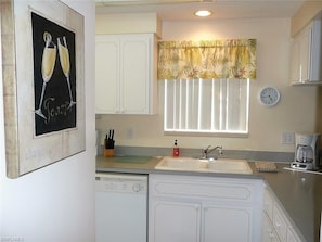 A kitchen with white cabinets, a sink, a dishwasher, a clock, and a window with blinds.
