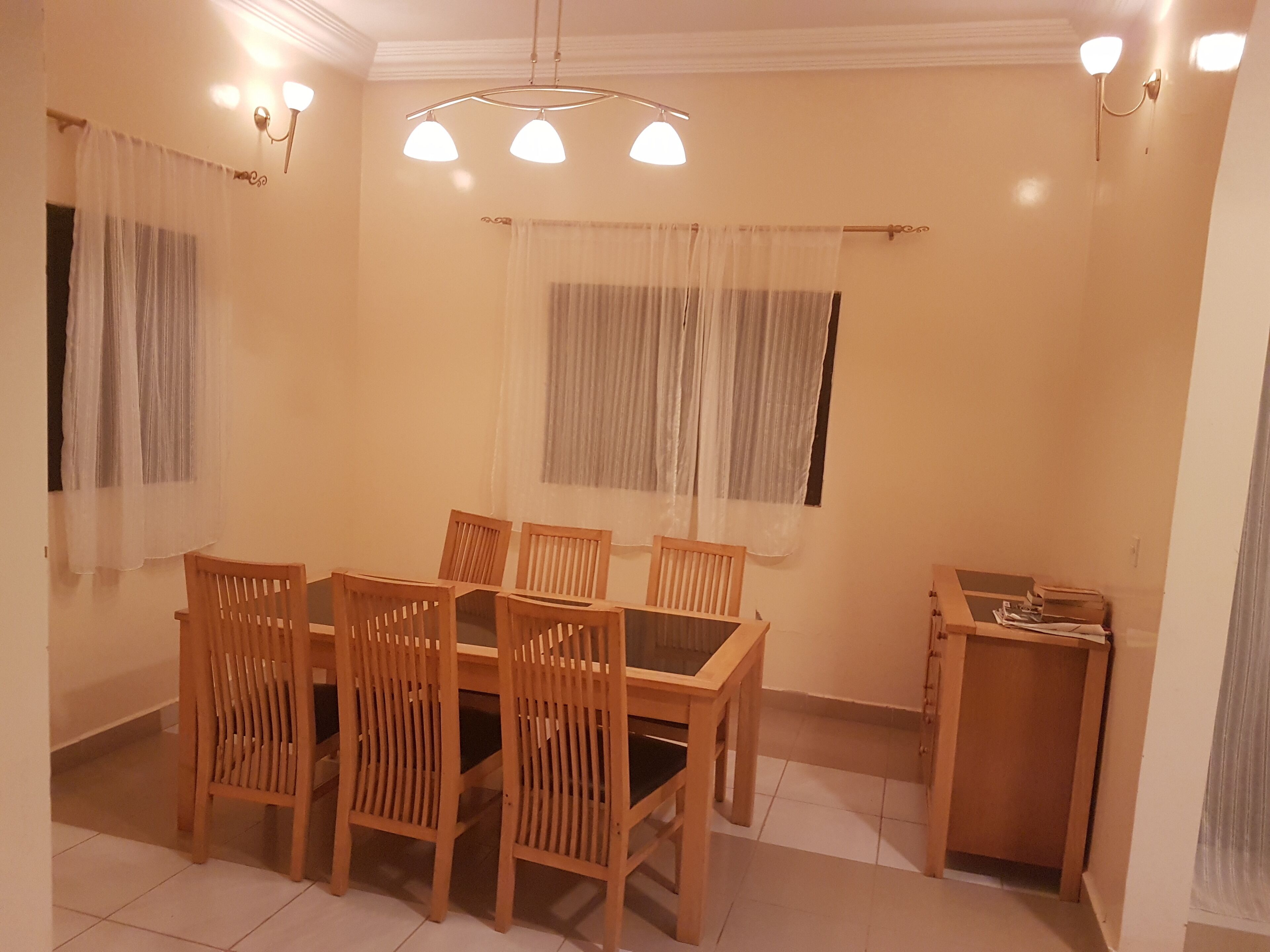 A dining area with a wooden table and chairs, a small cabinet, and two windows with curtains.