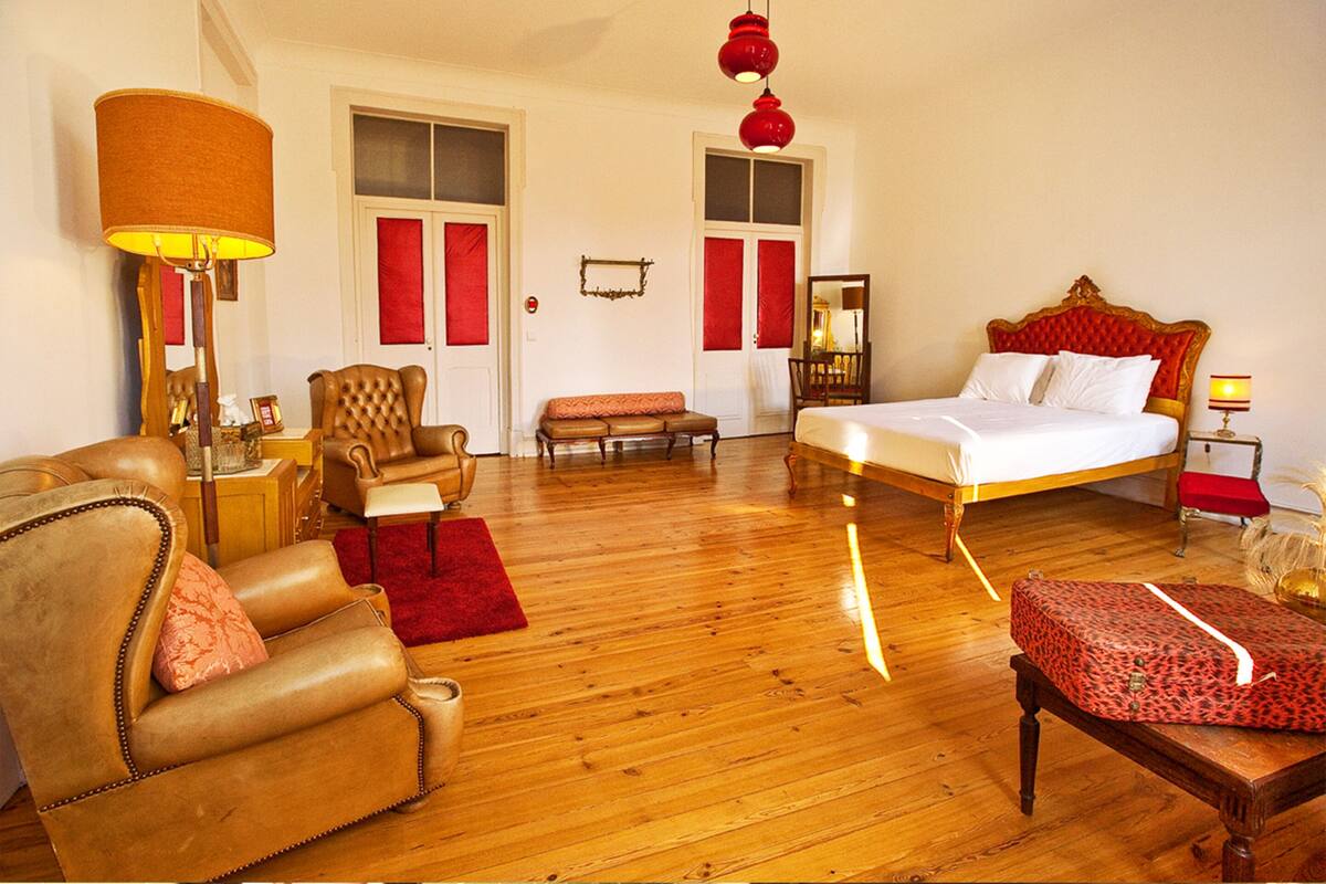 A spacious bedroom with a large bed, a leather armchair, a wooden bench, and a red lamp.