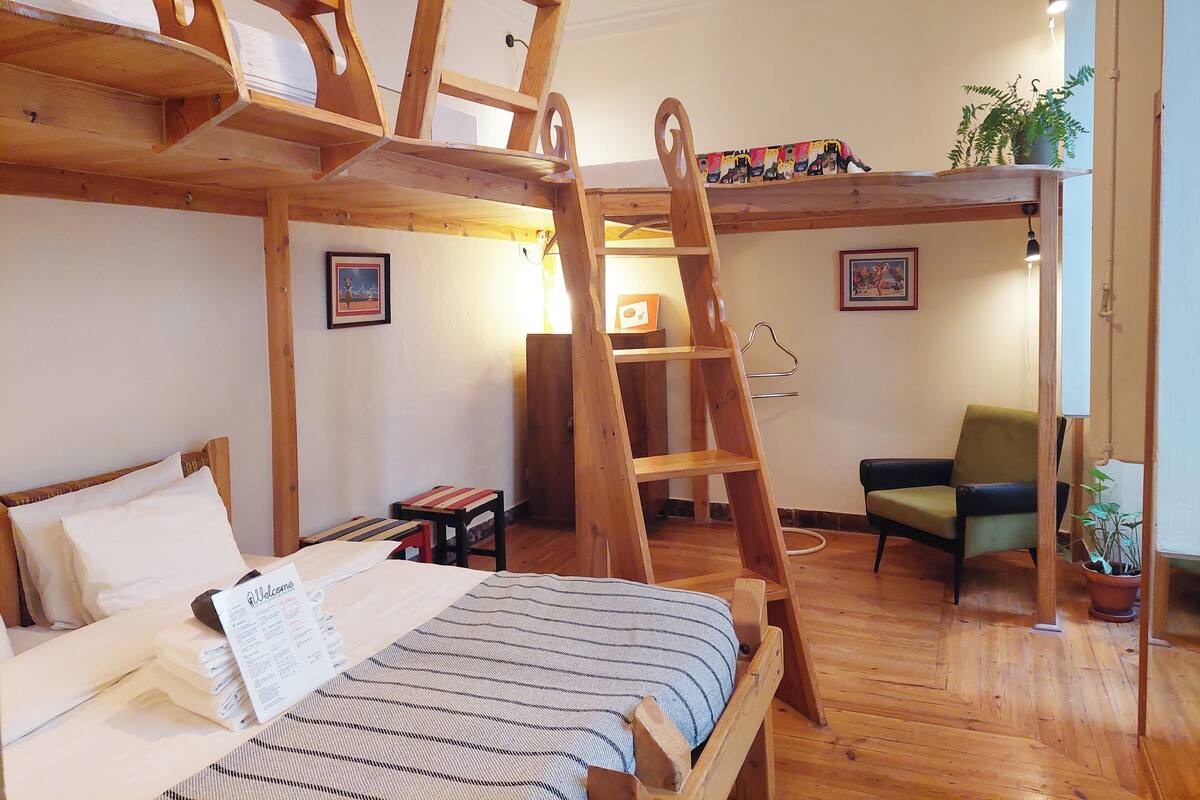 A wooden bunk bed with a ladder, a single bed with a striped blanket, a small table with a welcome sign, a green armchair, and a potted plant.
