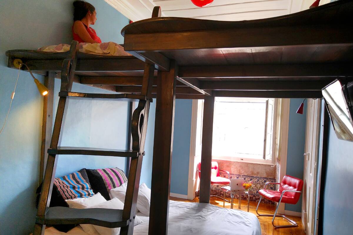 A bunk bed with a ladder, a desk with a laptop, and a red chair in a room with blue walls and a window.