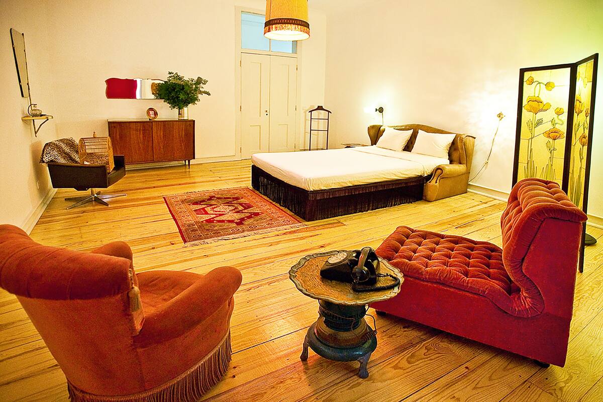A bedroom with a bed, a red sofa, a wooden floor, a small table with a telephone, a dresser, and a mirror.