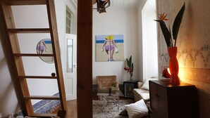 A room with a wooden ladder, a painting of a woman on the wall, a vase with flowers, and a cabinet. - Lisbon Calling (Lisbon)