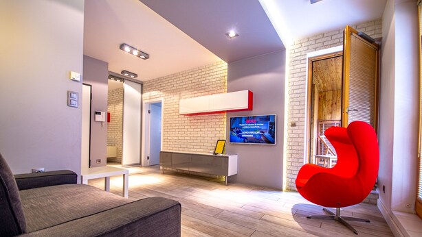A modern living room with a grey sofa, a red chair, a TV mounted on a brick wall, and a wooden floor.