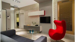 A modern living room with a grey sofa, a red chair, a glass coffee table, and a TV mounted on the wall.
