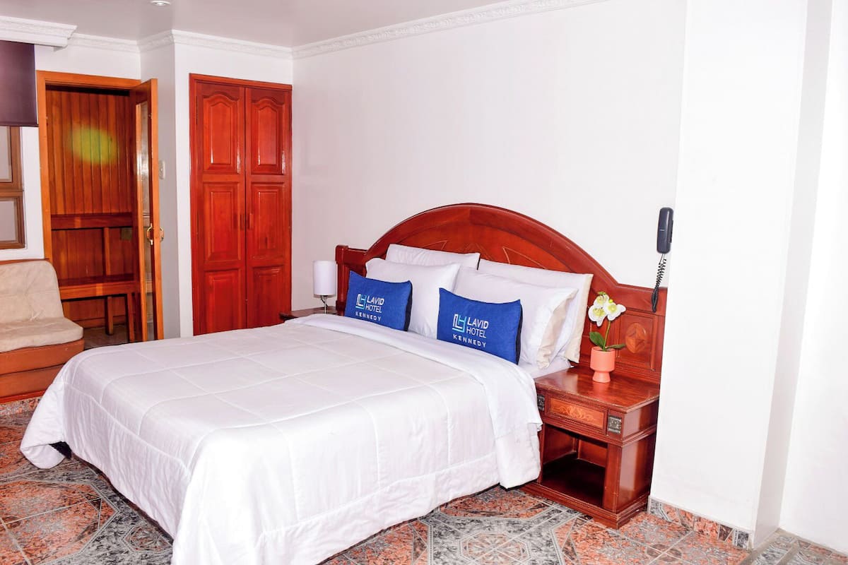 Une chambre d’hôtel avec une tête de lit en bois, un lit avec une literie blanche, deux oreillers bleus ornés du logo de l’hôtel, une table de chevet avec un téléphone et un petit vase avec une fleur.
