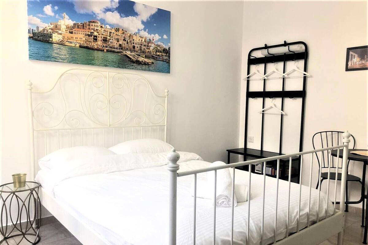 A neatly arranged bedroom with a white metal bed frame, a small bedside table, a chair, and a wall-mounted coat rack.