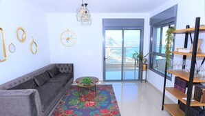 A modern living room with a grey sofa, a colorful rug, a small round table, and a bookshelf with books and glasses. - Domos Tiberias (Tiberias)