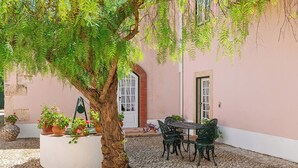 A courtyard with a tree, potted plants, a table with chairs, and a building with a door and window. - Casa da Avó Genoveva (Tomar)