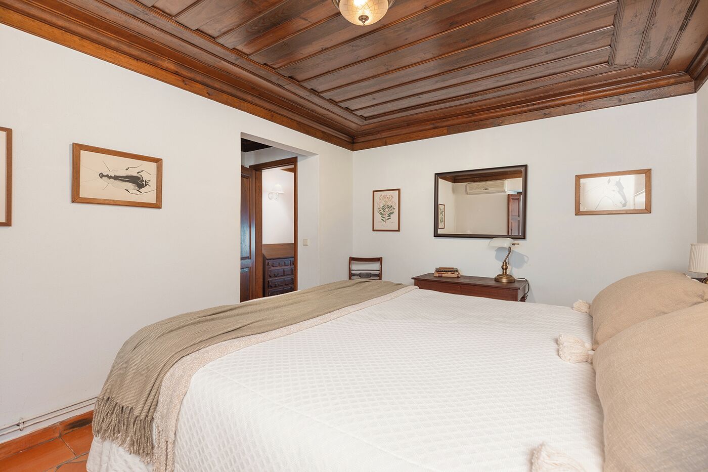 Un dormitorio con techo de madera, una cama con manta color beige, una mesita de noche con lámpara y cuadros enmarcados en las paredes.
