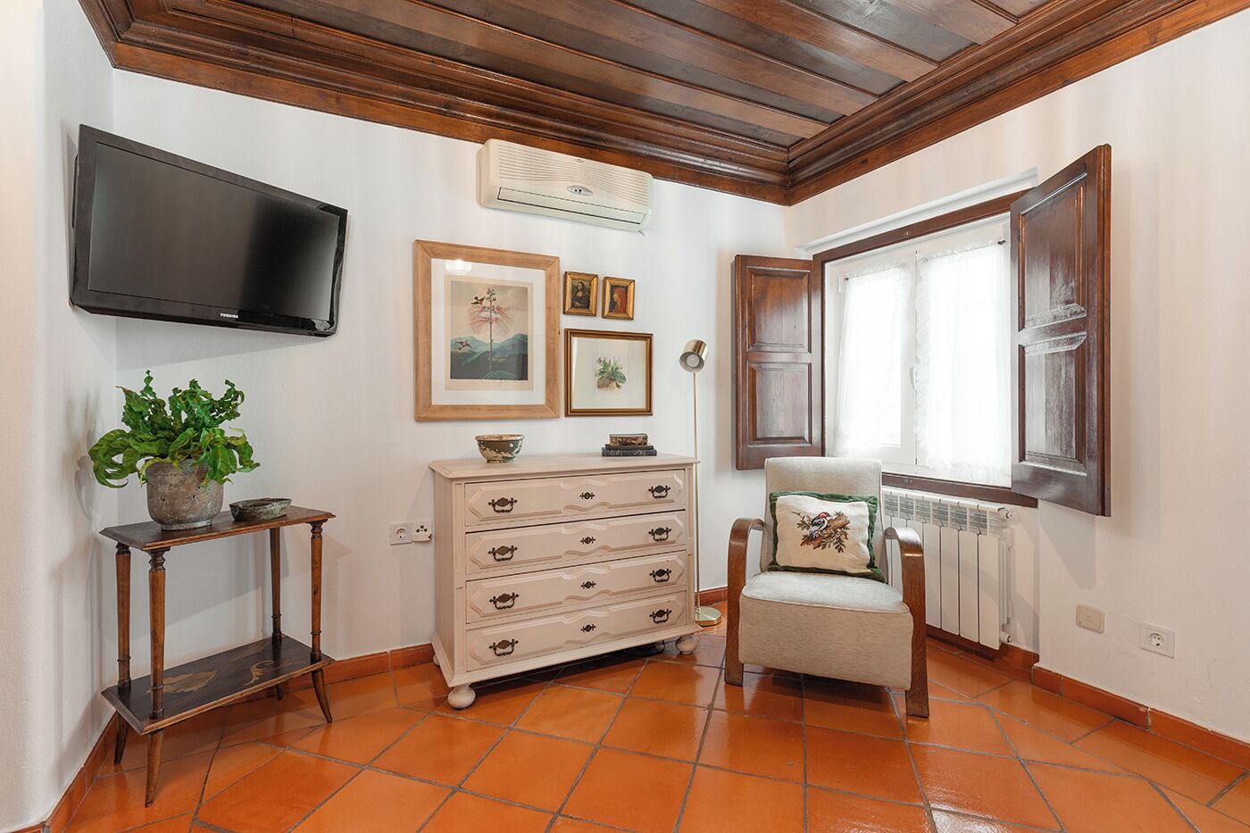 Quarto com teto de madeira, televisão fixada na parede, penteadeira com gavetas, cadeira com almofada, planta em vaso e janela com cortinas.