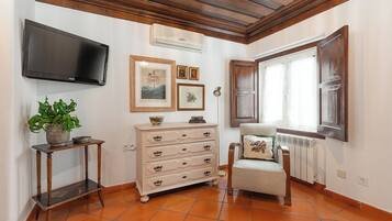 Quarto com teto de madeira, televisão fixada na parede, penteadeira com gavetas, cadeira com almofada, planta em vaso e janela com cortinas.