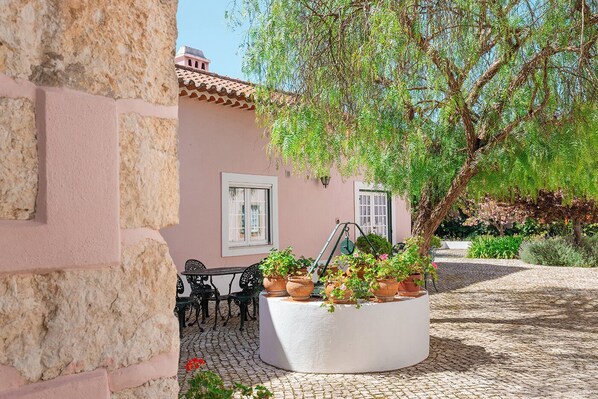 A courtyard with a white planter filled with potted plants, a table with chairs, and a building with a tiled roof.