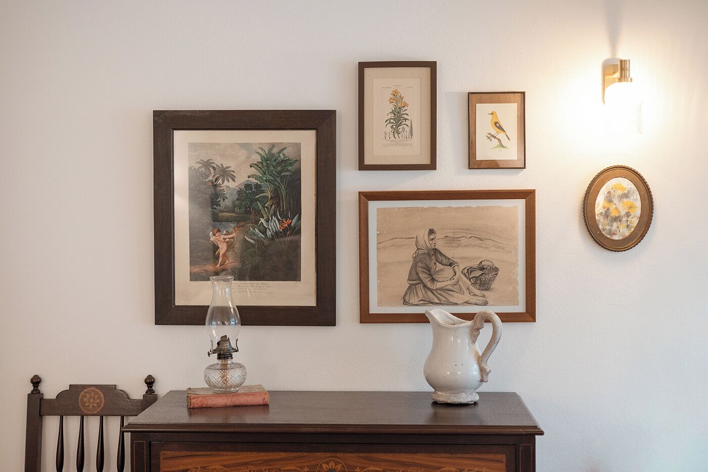En vägg med inramade botaniska illustrationer, en gammal kanna och en liten vas på ett träbord.